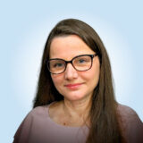 A person with long brown hair and glasses, wearing a light pink top, smiles softly at the camera against a light blue background.