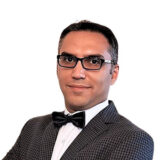 A person in a checkered suit and bow tie, wearing glasses, with a confident expression, against a plain white background.