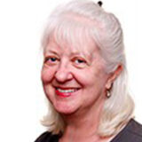 A smiling woman with white hair and earrings, wearing a gray top, stands against a plain white background, exuding warmth and friendliness.