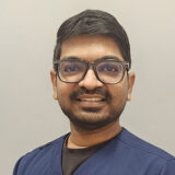 A friendly individual with glasses smiles in a professional attire, standing against a neutral background. The person appears approachable and confident, likely a healthcare or dental professional.