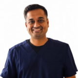 A smiling person wearing a dark blue scrub top stands against a plain white background.
