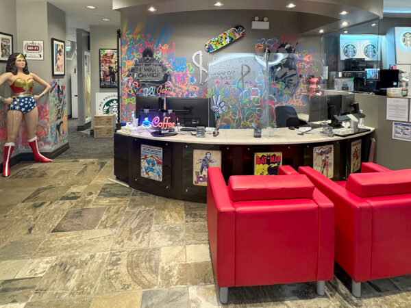 A modern reception area featuring a life-sized Wonder Woman figure, colorful wall art, and a stylish front desk. Two bright red armchairs are positioned in front of the desk, which is adorned with various colorful decorations and posters.