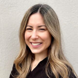 A smiling woman with long, wavy brown hair wearing a black top poses against a neutral background. Her bright smile conveys warmth and friendliness, creating an inviting atmosphere.