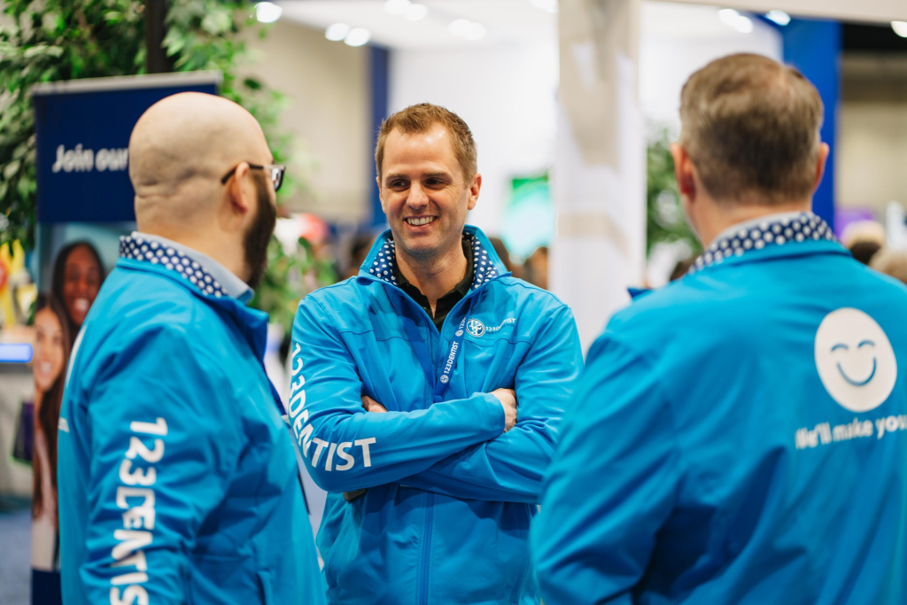 Three people in blue jackets are engaged in conversation at an event. One individual is smiling while the others face him. The jackets feature the 123Dentist branding, and there is a friendly blue smiley face visible on one of the jackets, adding a welcoming touch to the scene.