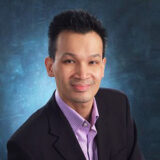A smiling man in a black suit and light purple shirt poses against a blue background, exuding professionalism and warmth.