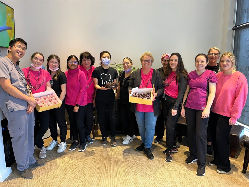 A group of dental professionals wearing various shades of pink stands together, smiling warmly. They are holding boxes of donuts, showcasing a festive atmosphere. A friendly blue smiley face, known as the 123Dentist Smiley, adds cheer to the gathering.