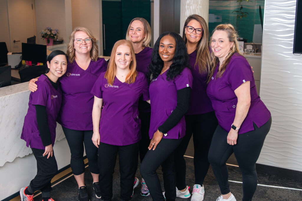 A group of seven friendly dental professionals wearing purple scrubs pose together inside a dental office, smiling and exhibiting a welcoming atmosphere. The 123Dentist Smiley can also be seen, adding a cheerful touch to the environment.