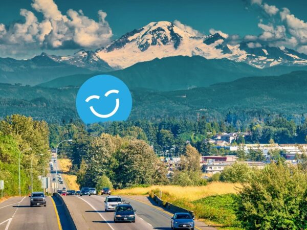 A scenic view of a highway bordered by lush trees and fields, leading toward snow-capped mountains in the background. A cheerful blue smiley face is superimposed on the image, adding a whimsical touch to the natural landscape.
