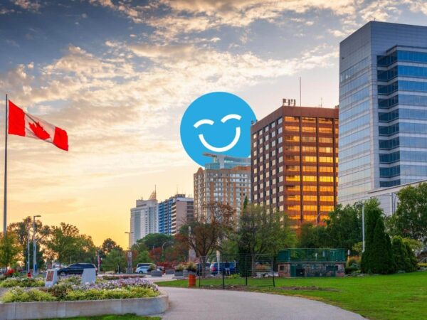 A city skyline at sunset featuring modern buildings and greenery, with a Canadian flag in the foreground and a smiling blue emoji overlay.