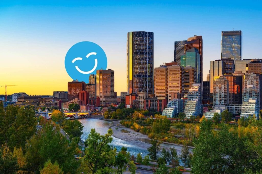 Skyline of a modern city at sunset with tall buildings, surrounded by greenery and a river; a large blue smiling emoji is overlaid on the scene.