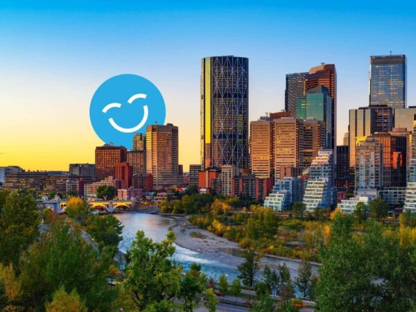 Skyline of a modern city at sunset with tall buildings, surrounded by greenery and a river; a large blue smiling emoji is overlaid on the scene.