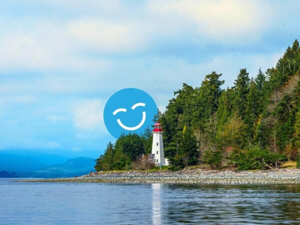 A lighthouse stands on a rocky shore surrounded by dense green trees, with calm water in the foreground and a partly cloudy blue sky. A blue, smiling emoji with closed eyes is superimposed in the sky.