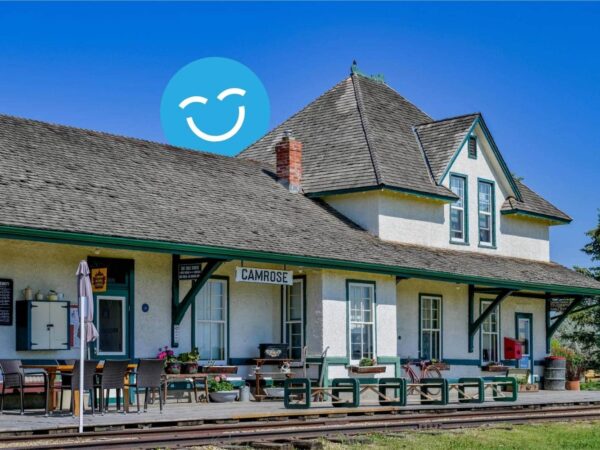 A historic train station with a sloped roof and wooden platform under a clear blue sky. A large blue circle with a smiling face is above the building.