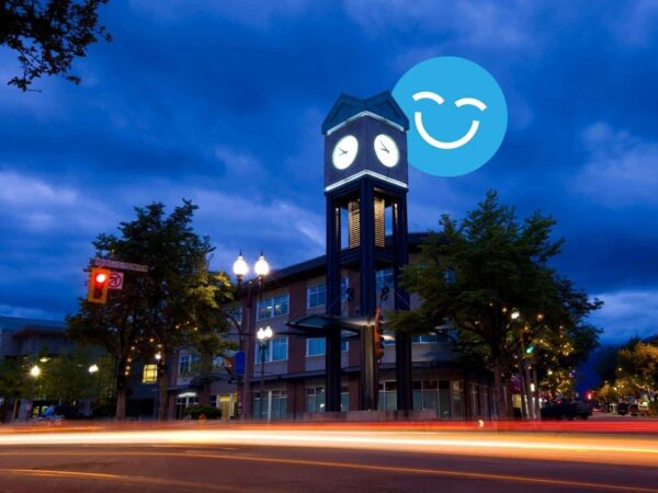 Clock tower illuminated at dusk with a smiling face in the sky, surrounded by trees and a street with light trails.