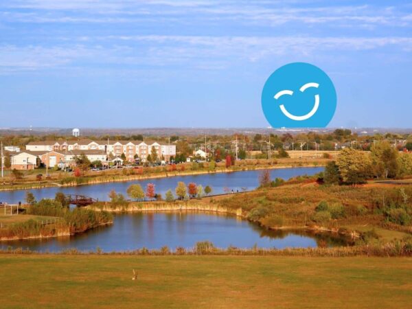 A peaceful landscape with calm water, autumn foliage, and a distant community. A blue smiley face symbol is prominent in the sky.