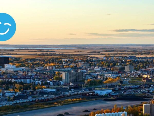 A scenic aerial view of a city at sunset, showcasing buildings and green spaces, with a river in the foreground and a soft, warm light illuminating the landscape. A blue smiley face icon appears in the corner.