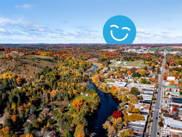Aerial view of a colorful autumn landscape featuring a winding river through vibrant foliage, with a small town visible along the riverbank. A cheerful blue smiley face icon is superimposed in the sky above.
