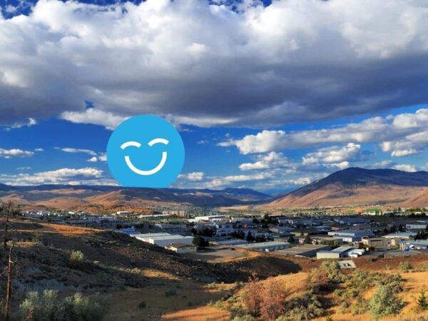 A panoramic view of a valley bordered by mountains under a partly cloudy sky. In the foreground, a mix of grassy and rocky terrain leads to a small industrial area. A blue smiley face icon overlays the image.