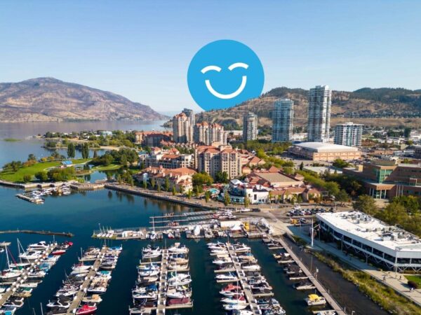Aerial view of a vibrant waterfront marina surrounded by modern buildings, with boats docked in the harbor and mountains in the background. A cheerful blue smiley face icon overlays the scene, adding a playful touch.