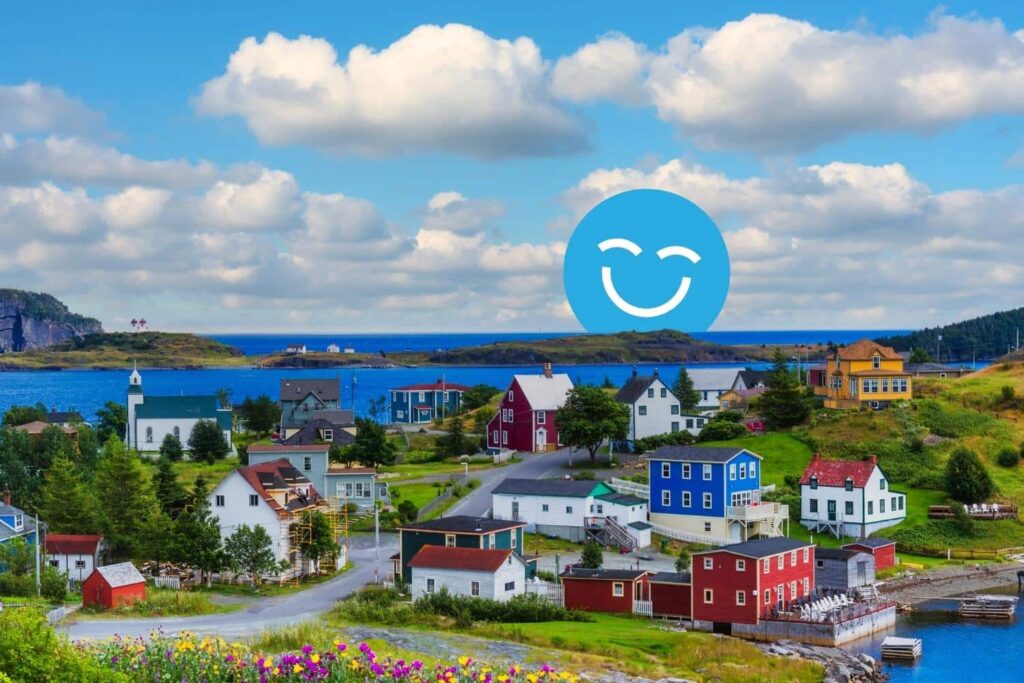 Un village côtier coloré présente diverses maisons à côté d'une mer bleue sereine sous un ciel lumineux avec des nuages duveteux. Un grand visage souriant se superpose à la scène, ajoutant un élément joyeux au paysage pittoresque. 