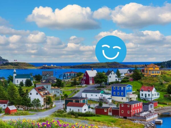 A colorful coastal village features various houses beside a serene blue sea under a bright sky with fluffy clouds. A large smiling face graphic overlays the scene, adding a cheerful element to the picturesque landscape.