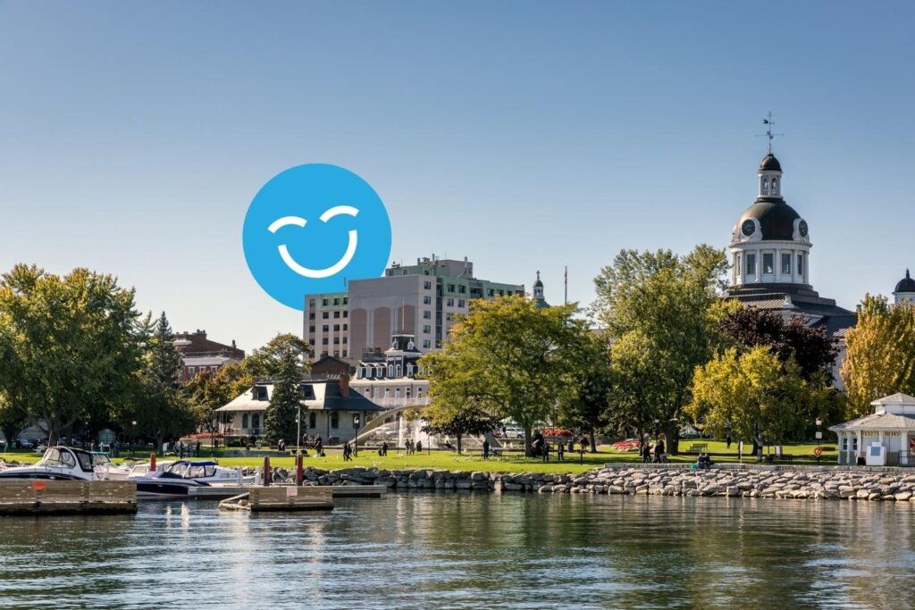 A serene waterfront scene with trees and a park, featuring buildings in the background and a large blue smiley face graphic in the sky. The calm water reflects the scenery, creating a peaceful atmosphere.