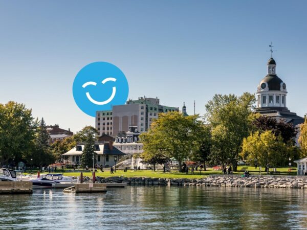 A serene waterfront scene with trees and a park, featuring buildings in the background and a large blue smiley face graphic in the sky. The calm water reflects the scenery, creating a peaceful atmosphere.