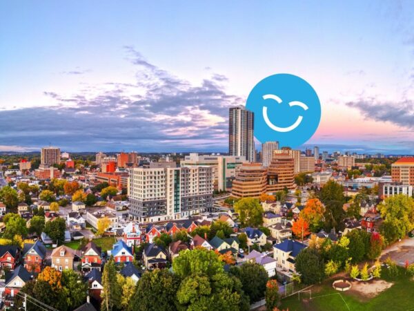 A picturesque urban skyline at sunset, showcasing a mix of modern buildings and residential areas. Vibrant fall foliage and a blue smiley face graphic superimposed enhance the scene, indicating a cheerful atmosphere.