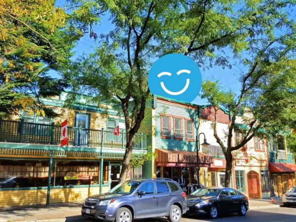 A vibrant street scene featuring colorful buildings with balconies, trees providing shade, and parked cars along the curb, all under a clear blue sky with a smiling face graphic overlay.