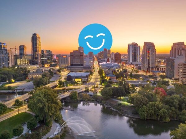 A scenic view of a city skyline at sunset, with tall buildings reflecting the warm colors of the sky. A river and green trees are in the foreground, and a smiling blue icon overlays the image.