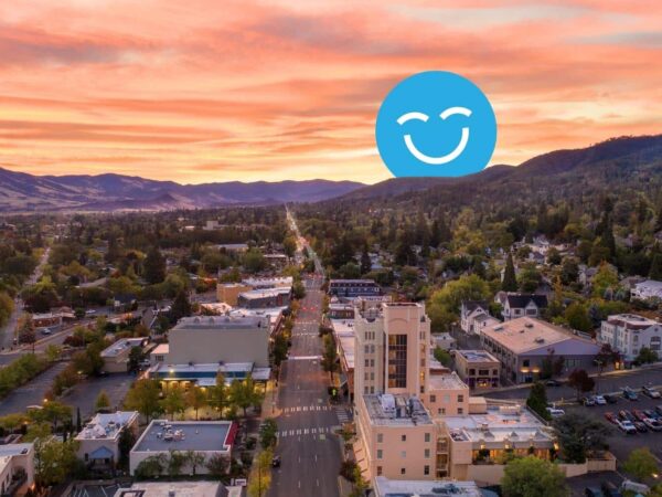 A scenic aerial view of a suburban town at sunset, with vibrant colors in the sky. A large, blue smiley face graphic is superimposed over the landscape, adding a whimsical touch to the serene setting.