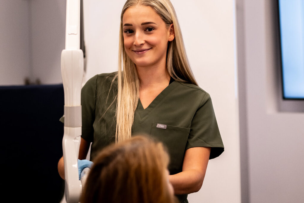 A dental professional with long blonde hair smiles while preparing for a procedure. She wears a dark green scrubs and is attentive to a patient seated in front of her. The setting appears to be a modern dental office.