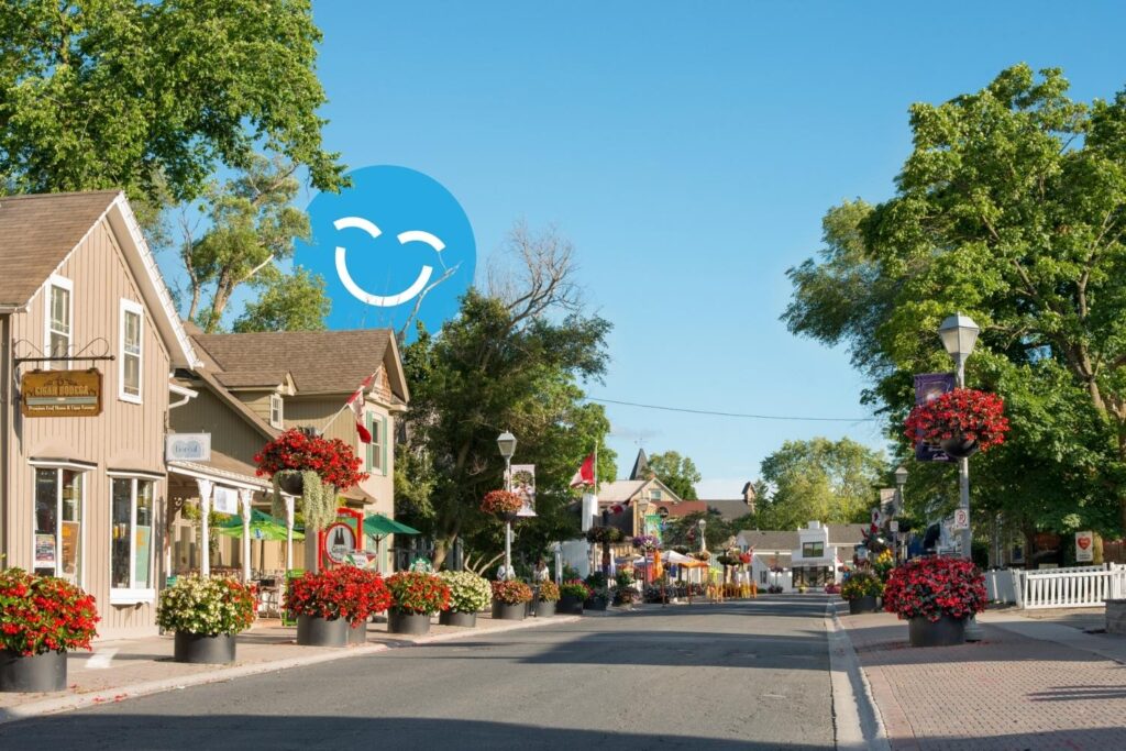 A quaint streetscape featuring charming buildings and vibrant flower arrangements, under a clear blue sky. A cheerful graphic smiley face is prominently displayed in the background, adding a playful touch to the serene scene.