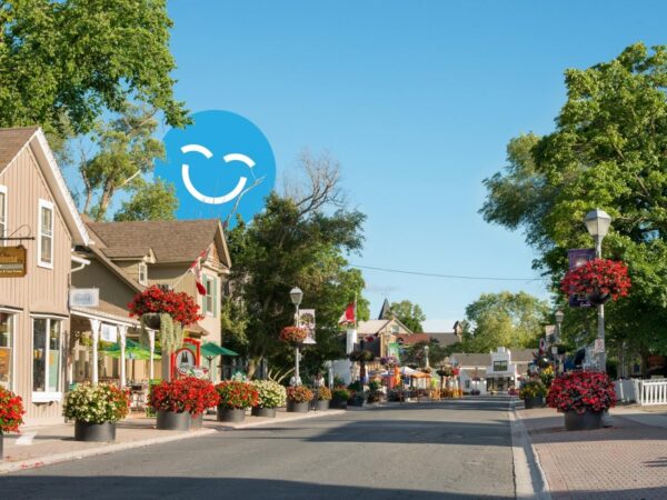 A quaint streetscape featuring charming buildings and vibrant flower arrangements, under a clear blue sky. A cheerful graphic smiley face is prominently displayed in the background, adding a playful touch to the serene scene.