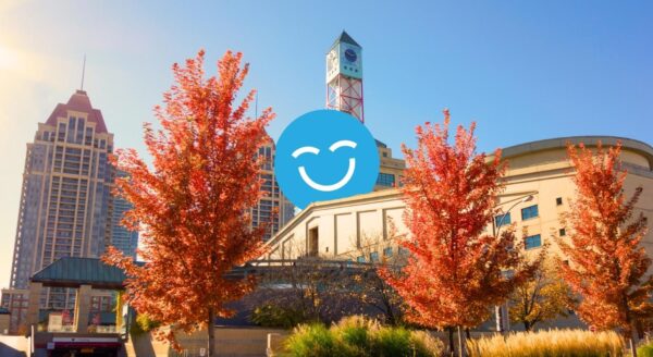 A sunny urban scene featuring modern buildings and vibrant red trees in autumn. A cheerful blue smiley face is superimposed over the landscape, adding a whimsical touch to the cityscape.