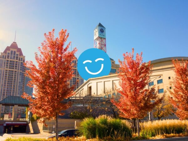 A sunny urban scene featuring modern buildings and vibrant red trees in autumn. A cheerful blue smiley face is superimposed over the landscape, adding a whimsical touch to the cityscape.