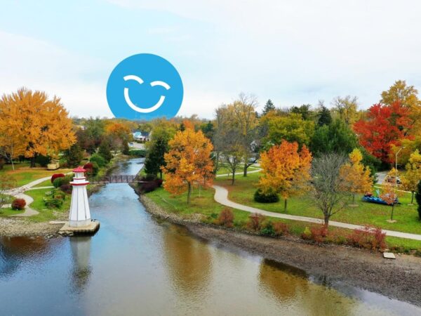 A serene park scene featuring vibrant autumn foliage, a calm river, and walking paths. A white lighthouse stands near the water, complemented by a playful blue smiley face in the background. The landscape captures the beauty of fall in a tranquil setting.
