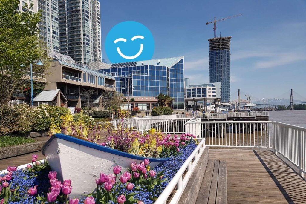 A waterfront scene features vibrant flowers in a wooden planter, with modern buildings in the background and a blue sky. A smiling face icon is superimposed above the scenery.