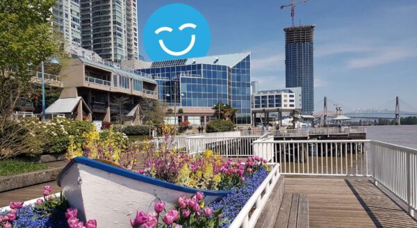 A waterfront scene features vibrant flowers in a wooden planter, with modern buildings in the background and a blue sky. A smiling face icon is superimposed above the scenery.