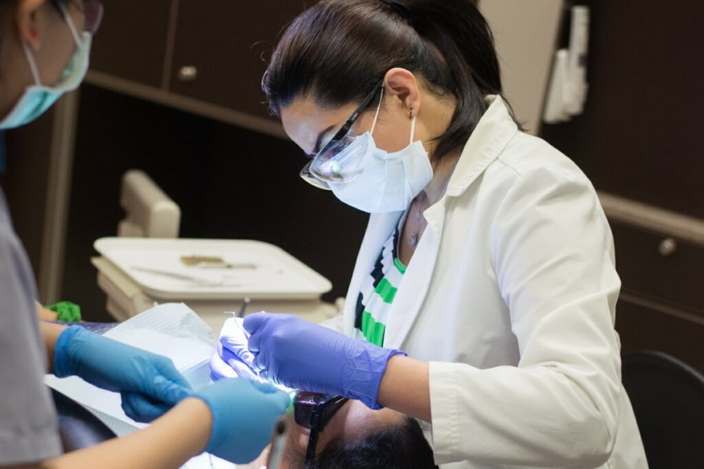 A dentist in a white lab coat performs an examination on a patient, while an assistant hands tools. Both wear masks and gloves, focused on providing dental care in a modern clinic setting. The friendly 123Dentist Smiley adds a cheerful ambiance.