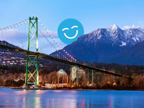A colorful suspension bridge spans a river, with snow-capped mountains in the background. A cheerful, smiling icon overlays the scene, adding a vibrant touch to the landscape at dusk.