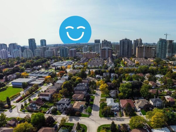 Aerial view of a suburban neighborhood with various houses, trees, and green spaces, contrasted by a skyline of urban buildings in the background. A large blue smiley face graphic is prominently displayed above the scene.