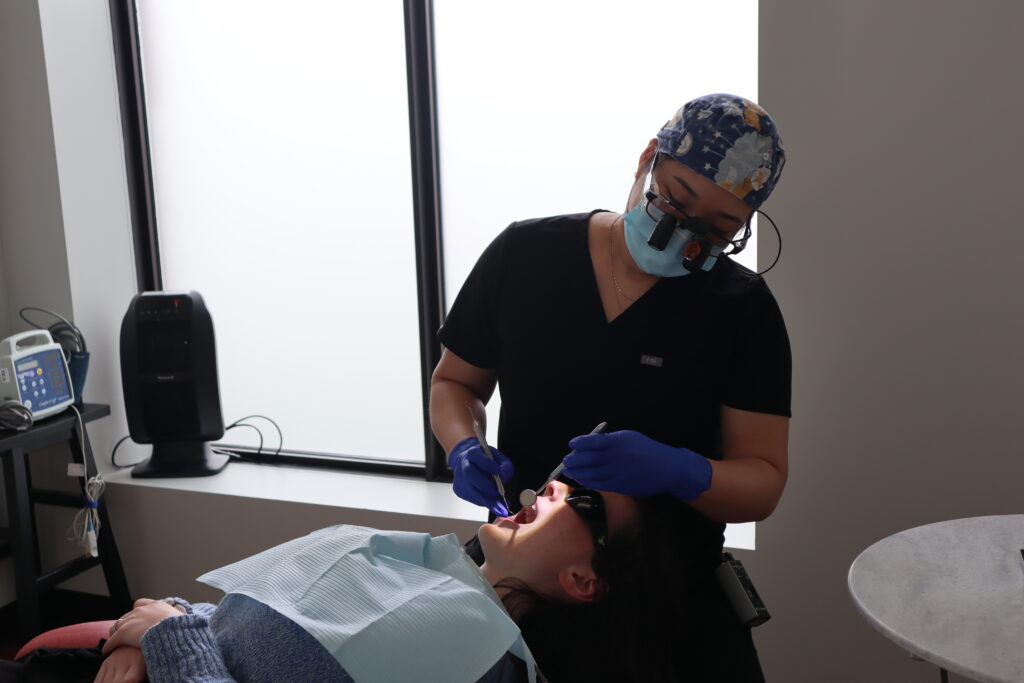 A dental professional, wearing scrubs and gloves, is providing treatment to a patient seated in a dental chair. The patient is wearing protective eyewear and has a dental bib on. Natural light enters the room through a large window.