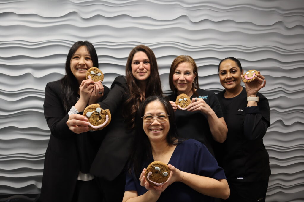 Five dental professionals smile while holding cookies in a dental office setting, showcasing a lighthearted atmosphere. The group includes a friendly 123Dentist Smiley, adding to the cheerful environment.