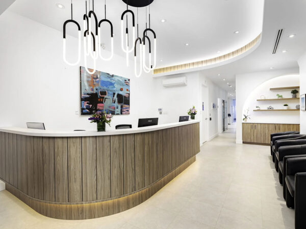 Modern dental clinic reception area with a curved wooden desk, contemporary pendant lights, a decorated wall, and a cozy waiting area with black chairs on the right.