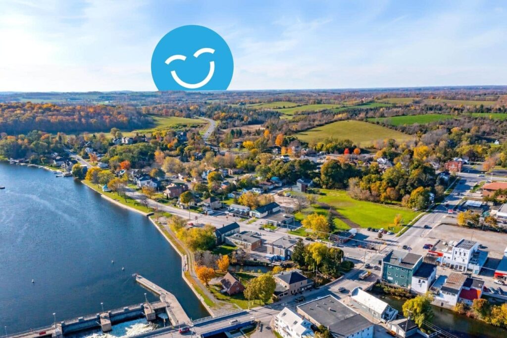 Une vue aérienne pittoresque d'une petite ville nichée au bord d'une rivière, entourée de feuillages d'automne colorés et de collines ondulantes. Un joyeux visage souriant bleu est superposé dans le ciel.