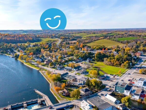 A scenic aerial view of a small town nestled beside a river, surrounded by colorful autumn foliage and rolling hills. A cheerful blue smiley face is overlaid in the sky.