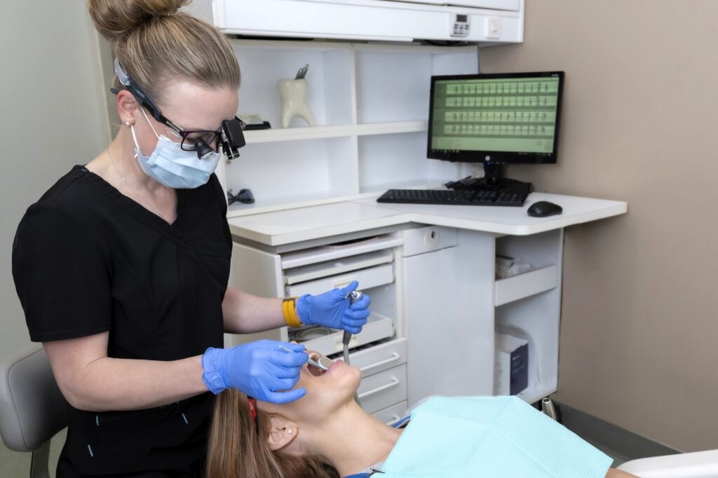 A dental hygienist wearing protective gear attends to a patient in a dental office, preparing for a procedure. The patient is reclined with a dental bib, while a computer workstation is visible in the background. The friendly 123Dentist Smiley adds a cheerful touch to the setting.