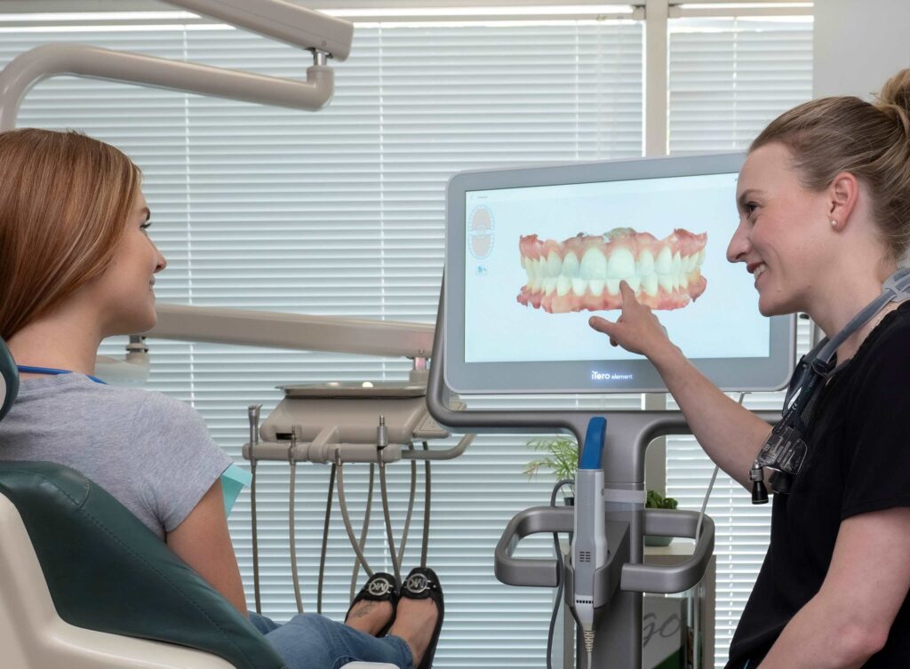 A dental professional explains a dental model on a screen to a patient seated in a dental chair. The setting conveys a friendly and informative atmosphere, focusing on patient care and education in a modern dental office.
