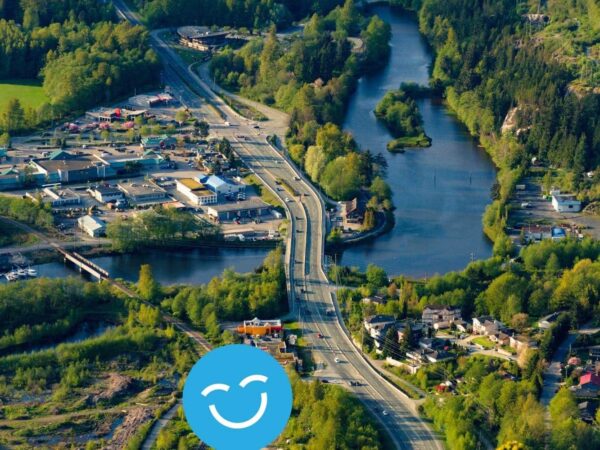 Aerial view of a lush landscape featuring winding roads, a river, and clusters of green trees. Nearby, residential and commercial buildings are visible, surrounded by nature. A cheerful graphic is superimposed at the bottom.